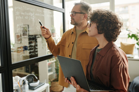 Young woman with laptop looking at notes on transparent board and listening to colleague explaining...