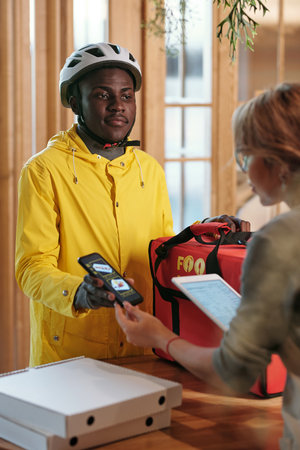 Young black man in uniform and waitress checking prices of packed online orders of clients while courier holding smartphone with advertの写真素材