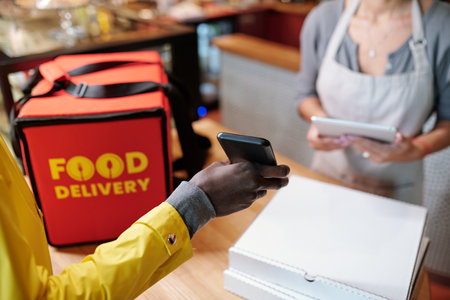 Hand of courier holding smartphone over table with big red bag and packed boxes with pizza and showing online menu items to waitressの写真素材