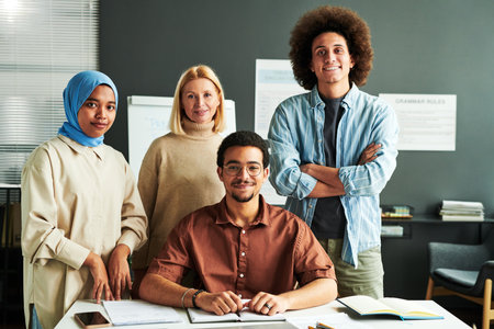 Group of multicultural students and mature blond teacher in casual attire standing by desk in front...