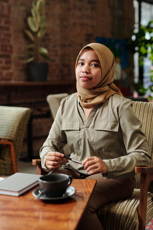 Young successful African American Muslim female employee or intern in hijab and casualwear sitting...