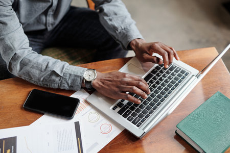 Above angle of hands of young African American businessman pressing buttons of laptop keyboard while...