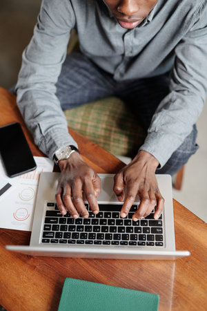 Above angle of young African American economist or analyst keeping hands over laptop keyboard while...
