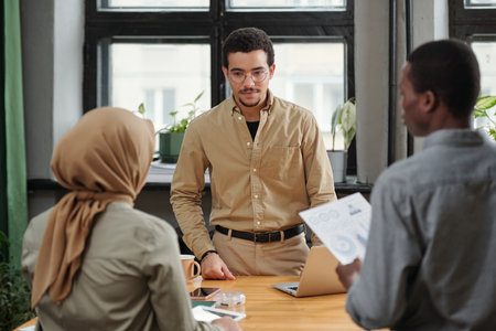 Young serious manager listening to report of Muslim female subordinate while standing in front of two intercultural employees at meetingの写真素材