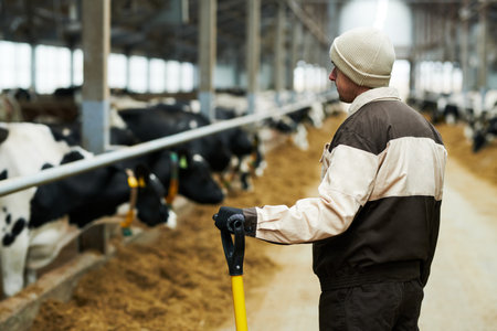 Back view of young male worker of cowfarm with worktool standing in front of camera and looking at...