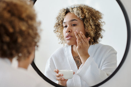 Young attractive woman applying moisturizing cream on face in front of mirror while taking care of her beauty after morning showerの写真素材