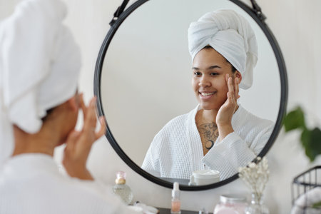 Happy young woman with towel on head applying moisturizing cream on face while looking in mirror in bathroom after morning showerの写真素材