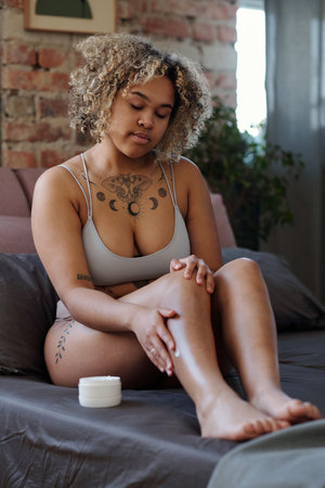 Young woman in underwear sitting on bed and applying hydrating lotion or cream on legs after shaving while taking care of her bodyの写真素材