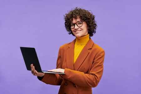 Happy young successful teacher with laptop looking at camera while standing in isolation on violet background during photo session in studioの写真素材