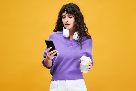 Cute female student in lavender v-shirt and while pants using mobile phone and having cup of tea at...