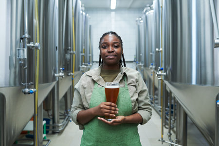 Portrait of positive female worker in uniform showing glass of beer produced in her breweryの写真素材