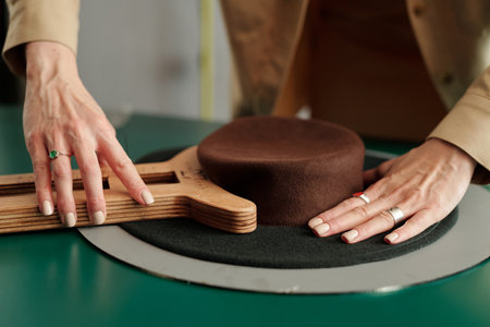 Hands of young female designer forming shape of brown felt porkpie hat with wooden tool by workplace...