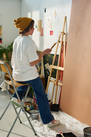 Young creative woman in stylish casualwear sitting in front of easel and huge panel by wall for painting new masterpieces in home studioの写真素材