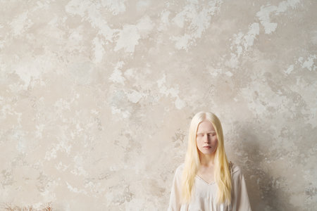 Young serene albino woman in white pullover keeping her eyes closed while standing by fair marble wall with copyspace on the leftの写真素材