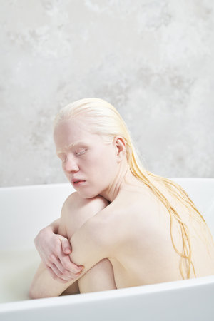 Peaceful albino girl with wet long hair sitting in warm water in white bathtub and embracing herself...