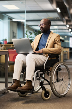 Young serious male analyst wearing casualwear with laptop on his knees sitting in wheelchair and networking in openspace officeの写真素材