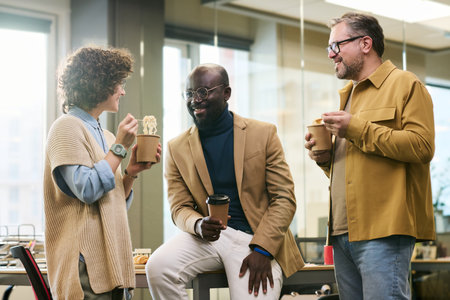 Happy young African American businessman with cup of coffee talking to colleagues eating Chinese wok at lunch break in openspace officeの写真素材