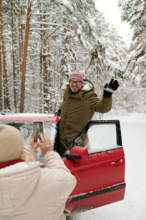 Cheerful mature male hiker in winterwear waving hand while standing in door of red car and posing...
