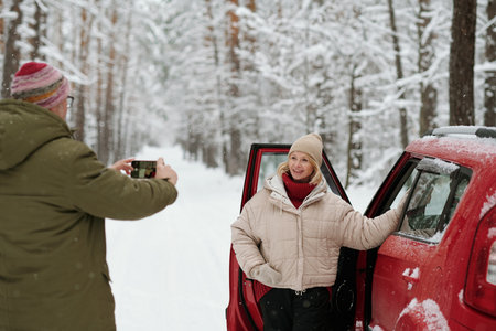 Smiling mature woman in winterwear standing by open door of car and posing for her husband with...