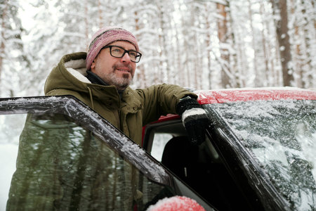 Smiling mature man in eyeglasses and winterwear standing by door of his car and enjoying winter vacation in the forest covered with snowの写真素材