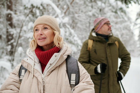 Senior blond woman in winterwear looking forwards during weekend stroll with her husband in pinetree forest covered with snowの写真素材