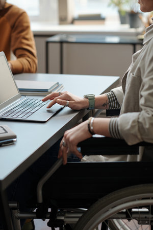 Close-up of young female manager with disability using laptop while sitting in wheelchair by workplace and analyzing online dataの写真素材