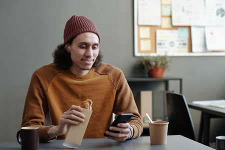 Young smiling multi-ethnic male employee with smartphone having sandwich and communicating in video...