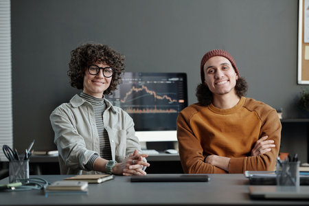 Two young intercultural managers in casualwear sitting by workplace in office and looking at camera against computer screen with graphの写真素材