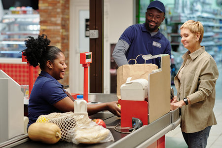 Happy young African American female cashier scanning food products chosen by mature customer standing in front of her in supermarketの写真素材