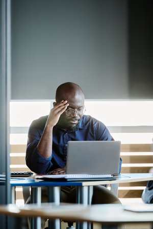 Serious young African American ceo sitting in front of laptop in office and communicating with...