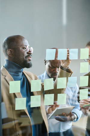 Young confident businessman in casualwear writing down tasks on notepapers while standing in front of transparent boardの写真素材