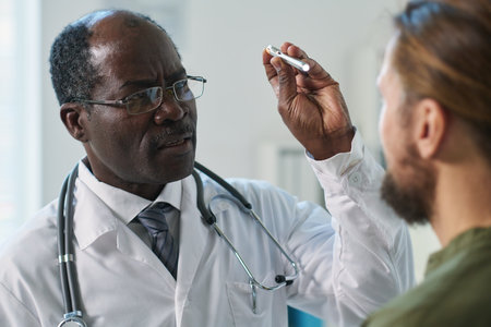 Mature male clinician with special medical device directing light on eyes of patient while checking...
