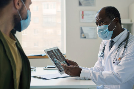 Confident mature radiologist in mask pointing at brain scan on tablet screen and looking at patient while explaining him exam resultsの写真素材
