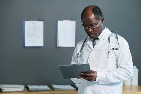 Serious mature general practitioner in lab coat making medical notes or prescriptions for patient in document while working in clinicsの写真素材