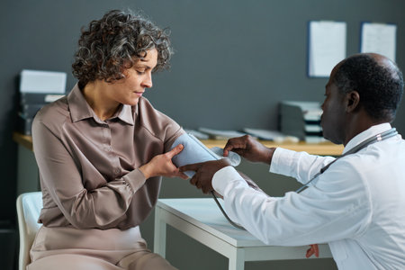 African American male doctor using tonometer while sitting by workplace in hospital and measuring blood pressure of mature female patientの写真素材