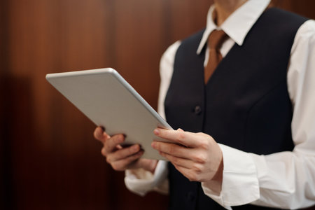 Close-up of young female staff of hotel in uniform using tablet by workplace while looking through new guests registered onlineの写真素材