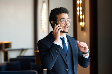 Confident young male entrepreneur in elegant dark blue suit talking to business partner on mobile phone in lounge of hotelの写真素材