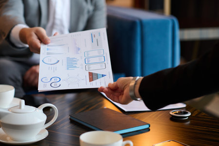 Close-up of financial document with graphic data being passed by analyst to colleague over table...