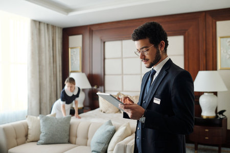 Young elegant entrepreneur in formalwear using tablet while standing in front of camera in hotel room with chambermaid on backgroundの写真素材