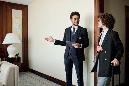 Young confident head manager of luxurious hotel showing room to female guest in smart casualwear with suitcase listening to himの写真素材