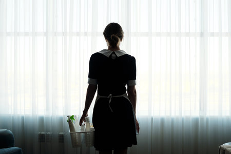 Rear view of chambermaid with detergents in basket walking towards large window covered with white chiffon curtains in hotel roomの写真素材
