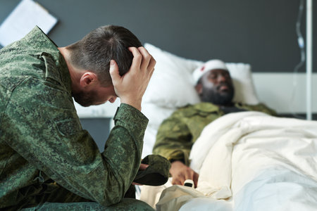 Young worried soldier in camouflage touching head while sitting by his friend in hospital bed after injury during military activitiesの写真素材