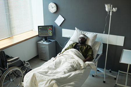 Young African American injured man in camouflage lying in bed between dropper and computer screen with medical indicatorsの写真素材