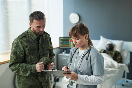 Young soldier in military uniform standing by doctor with medical document making notes or prescription for injured patientの写真素材