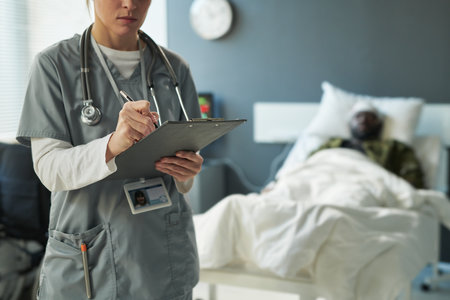 Close-up of young nurse in medical scrubs making medical prescriptions in document while standing against injured soldier lying in bedの写真素材
