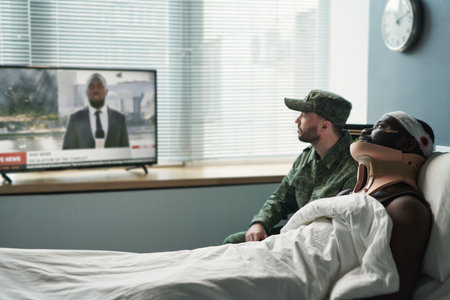 Side view of injured man and his buddy in military uniform watching news broadcast on tv while...
