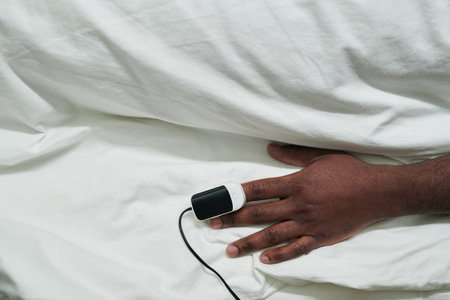 Hand of young African American male patient with pulse oximeter on fingertip lying under white cotton sheet in bed in hospital wardの写真素材