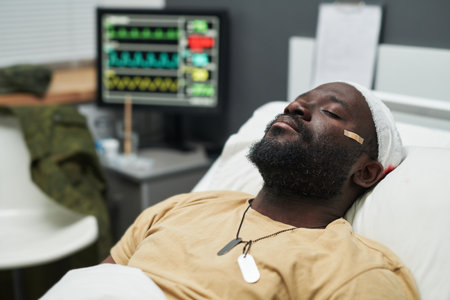 Close-up of young injured soldier with id tag on neck and bandage around his head lying in bed in hospital ward of intensive careの写真素材