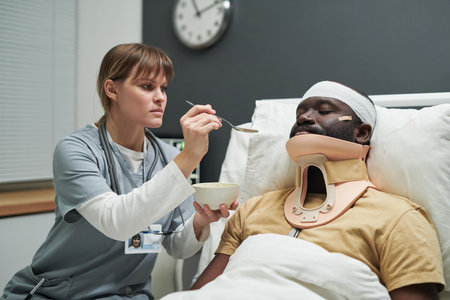 Nurse in uniform holding bowl with porridge while feeding injured patient in bandaged head and...
