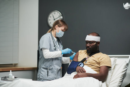 Nurse in uniform and protective mask making injection to wounded soldier with bandaged head and broken arm sitting in bed in hospital wardの写真素材